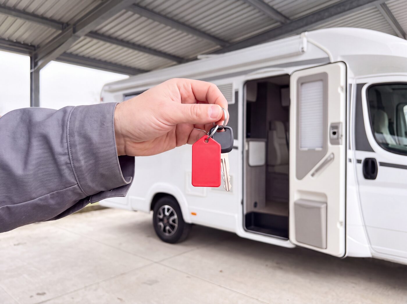 Una mano porge la chiave di un camper con la targhetta rossa davanti alla porta aperta del camper, sotto la tettoia del rimessaggio